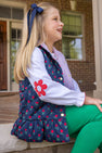 A young girl wears an Emma’s Elbow Patch Top - Worth Avenue White with Richmond Red Flowers under a navy blue quilted vest with a red tulip print and bright green pants.