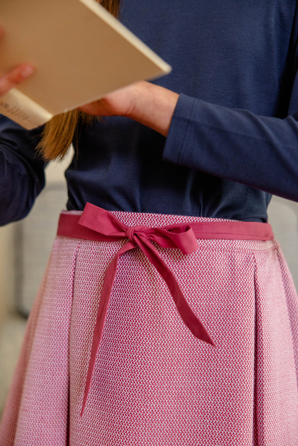 A person wearing a navy blue long-sleeve top and The Beaufort Bonnet Company’s Parson Pleated Skirt in Begley Berry Tweed, which features a bow at the waist, is holding an open book, with the focus on the outfit and hands.