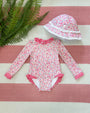 A Sarasota Surf Suit (UPF 35+) in the Mableton Mini Floral (Pink) with Romany Rose print from The Beaufort Bonnet Company, featuring long sleeves, delicate lace trim, and an adorable matching floral sunhat with white lace, is displayed on a pink and white striped surface beside green fern leaves.