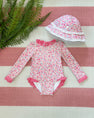 A Sarasota Surf Suit (UPF 35+) in the Mableton Mini Floral (Pink) with Romany Rose print from The Beaufort Bonnet Company, featuring long sleeves, delicate lace trim, and an adorable matching floral sunhat with white lace, is displayed on a pink and white striped surface beside green fern leaves.