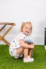 A smiling toddler with blonde hair and a white bow squats on green grass while holding a wooden toy. She wears The Beaufort Bonnet Company’s Michigan Britt Bubble in Bold in Blue with Hamptons Hot Pink. A small stool and part of another object are visible nearby.