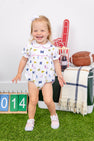 A smiling toddler stands on artificial grass wearing The Beaufort Bonnet Company’s Michigan Britt Bubble - Bold in Blue with Hamptons Hot Pink, crafted from soft Pima Cotton Knit. Behind her are a football, a foam finger, a striped blanket, a drink, and a 2014 sign. She pairs her outfit with pink shoes and a white bow.