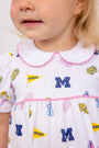A young child wears The Beaufort Bonnet Company’s Michigan Britt Bubble - Bold in Blue with Hamptons Hot Pink, a soft Pima Cotton Knit dress adorned with blue Ms, yellow Michigan pennants, and blue and yellow pom-poms. The dress features pink trim and a white Peter Pan collar, with only the child’s lower face visible.