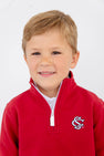 A young boy with short blond hair is smiling in front of a plain white background, wearing The Beaufort Bonnet Company’s USC Hayword Half-Zip in South Carolina Burgundy with Worth Avenue White.
