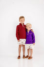 Two young boys stand together, smiling. One wears The Beaufort Bonnet Company’s USC Hayword Half-Zip in South Carolina Garnet with Worth Avenue White, paired with matching shorts in Gamecock style. The other wears a purple pullover and shorts featuring LSU colors. Both have light brown shoes and stand against a plain white background.