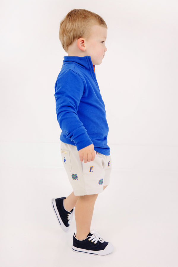 A young child in a blue zip-up sweater and white sneakers stands sideways against a plain white background, wearing The Beaufort Bonnet Company’s Florida Critter Sheffield Shorts in Saratoga Stone—perfect kids’ gameday shorts featuring fun Florida logo details.