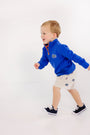 A young child wearing The Beaufort Bonnet Company’s Florida Hayword Half-Zip in Florida Blue with Florida Green & Orange and beige shorts walks happily in sneakers against a white background.