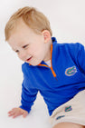 A young child with light hair is smiling while wearing The Beaufort Bonnet Company’s Florida Hayword Half-Zip in vibrant Florida Blue with Florida Green and Orange accents, featuring an orange zipper and the University of Florida Gators logo, paired with matching shorts, against a white background.