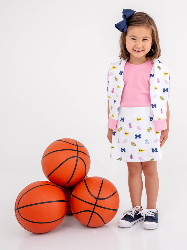 A young girl beams beside a stack of four basketballs, wearing The Beaufort Bonnet Company’s Michigan Cassidy Comfy Crewneck in Bold in Blue with Hamptons Hot Pink. She completes her spirited look with a navy hair bow and stands against a plain white background.