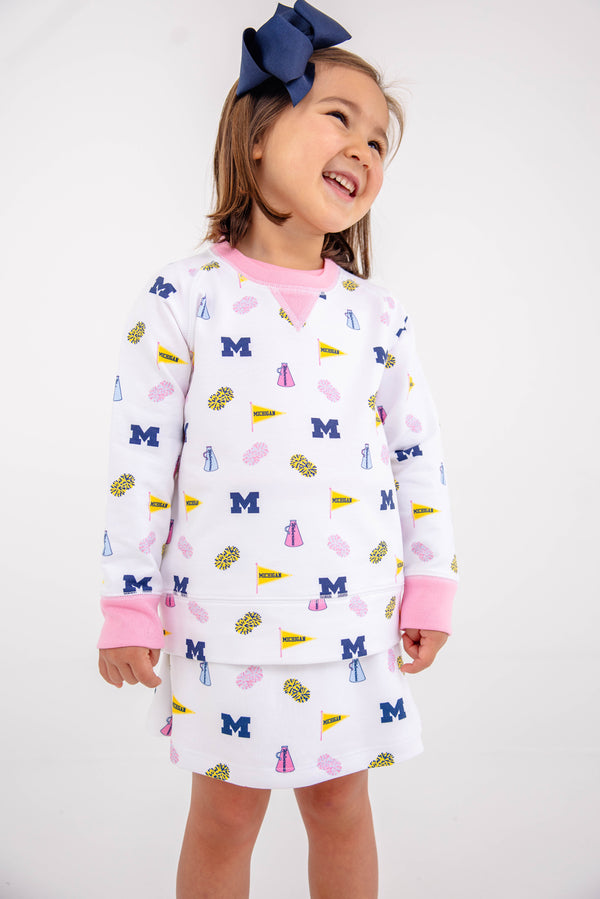 A young girl smiles, dressed in The Beaufort Bonnet Company’s Michigan Searcy Skort - Bold in Blue with Worth Avenue White. Her white outfit features pink cuffs and a playful pattern of blue Ms, pink dresses, yellow flags, and pom-poms. She wears a large navy blue bow in her hair and stands against a plain white background in this soft Pima cotton skort.