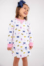 A young girl smiles, dressed in The Beaufort Bonnet Company’s Michigan Searcy Skort - Bold in Blue with Worth Avenue White. Her white outfit features pink cuffs and a playful pattern of blue Ms, pink dresses, yellow flags, and pom-poms. She wears a large navy blue bow in her hair and stands against a plain white background in this soft Pima cotton skort.