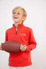 A smiling young boy wears The Beaufort Bonnet Company’s Ole Miss Hayword Half-Zip in Ole Miss Red with Nantucket Navy, featuring an embroidered logo, while holding a football with both hands—perfect for game day layering. The background is plain white.