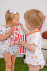 Two toddlers in patterned outfits, one dressed in The Beaufort Bonnet Company’s Tennessee Short Sleeve Patton Play Bubble – Victory in Orange with Tennessee Orange, share popcorn from red and white striped boxes. One child feeds popcorn to the other as they stand on green grass with a football and cooler in the background.