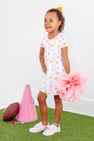 A young girl wearing the Tennessee Polly Play Dress - Victory in Orange with Palm Beach Pink from The Beaufort Bonnet Company, featuring cheerful patterns on soft Pima Cotton Knit, stands smiling while holding a large pink pom-pom. Beside her on the grass, against a white wall, lie a pink megaphone and a football.