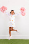 A smiling young girl jumps in the air holding pink pom-poms, wearing The Beaufort Bonnet Company’s Tennessee Searcy Skort - Victory in Orange with Worth Avenue White, paired with a white top featuring colorful patterns and pink sneakers. She stands on green artificial grass against a plain white background.