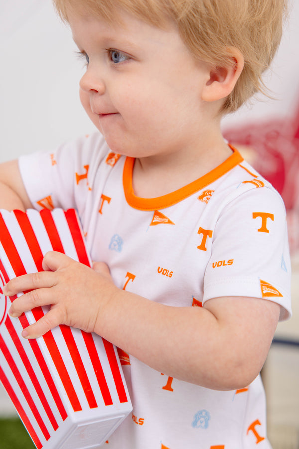 A toddler with light hair holds a red and white striped container. The child is dressed in The Beaufort Bonnet Company’s Tennessee Short Sleeve Patton Play Bubble – Victory in Orange with Tennessee Orange, featuring orange trim and symbols like “VOLS,” pennants, and the letter “T.”.
