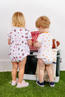 Two toddlers stand on green turf facing away, dressed in The Beaufort Bonnet Company’s Tennessee Short Sleeve Patton Play Bubble – Victory in Orange with Tennessee Orange. They stand near a cooler with a football, a blanket, and a popcorn container, creating a playful, sporty outdoor atmosphere.
