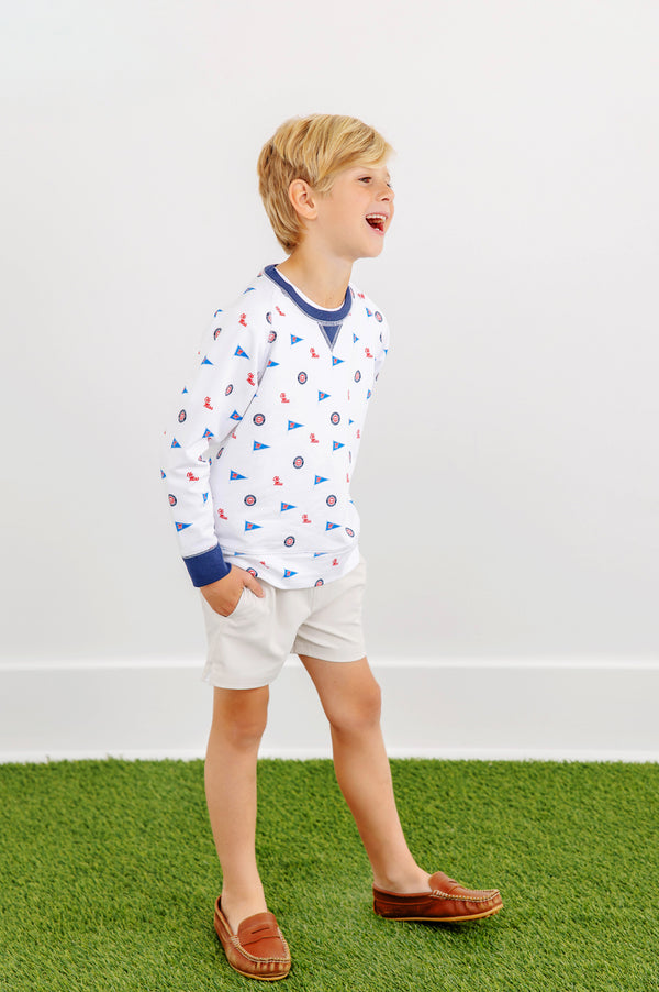 A young boy with blond hair, dressed in a white patterned long-sleeve shirt, khaki shorts, and brown loafers, stands on green artificial grass and smiles while looking to the side against a plain white background. He completes his look with The Beaufort Bonnet Company’s Ole Miss Cassidy Comfy Crewneck - Gawsh Almighty with Nantucket Navy for a fun and casual style.