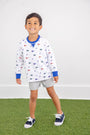 A young boy smiles while wearing The Beaufort Bonnet Company’s Florida Cassidy Comfy Crewneck – Florida Forever with Florida Blue, featuring colorful flag patterns, paired with gray shorts and navy sneakers, standing on green artificial grass against a plain white wall.