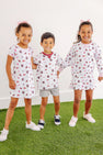 Three smiling young children in white sports-themed outfits stand on green turf against a white wall, holding hands. Their looks feature the South Carolina Spirit: two girls wear hair bows and The Beaufort Bonnet Company’s USC Cassidy Comfy Crewneck – South Carolina Spirit with South Carolina Burgundy—one styled with a dress and the other with a skirt—while the boy wears shorts.