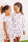 Two young girls with curly hair and pink bows smile at each other, both wearing matching USC Polly Play Dresses – South Carolina Spirit with Palm Beach Pink by The Beaufort Bonnet Company. Their soft Pima cotton outfits feature various pink and maroon logos and patterns inspired by South Carolina Spirit, as they stand against a plain white background.
