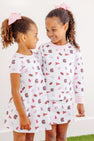 Two young girls with curly hair and pink bows smile at each other, both wearing matching USC Polly Play Dresses – South Carolina Spirit with Palm Beach Pink by The Beaufort Bonnet Company. Their soft Pima cotton outfits feature various pink and maroon logos and patterns inspired by South Carolina Spirit, as they stand against a plain white background.