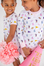 Two young girls smile at each other, wearing matching LSU Polly Play Dresses in the Tenacious Tigers with Palm Beach Pink print by The Beaufort Bonnet Company. One holds pink pom-poms, while the other carries a pink megaphone that says TIBERI.