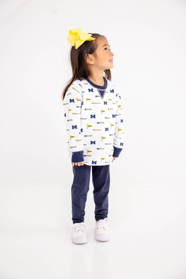 A young girl with a big yellow bow stands smiling and looking to the side against a plain white background, wearing The Beaufort Bonnet Company’s Michigan Cassidy Comfy Crewneck – Bold in Blue with Michigan Blue, paired with navy pants and white sneakers.