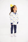 A young girl with a big yellow bow stands smiling and looking to the side against a plain white background, wearing The Beaufort Bonnet Company’s Michigan Cassidy Comfy Crewneck – Bold in Blue with Michigan Blue, paired with navy pants and white sneakers.