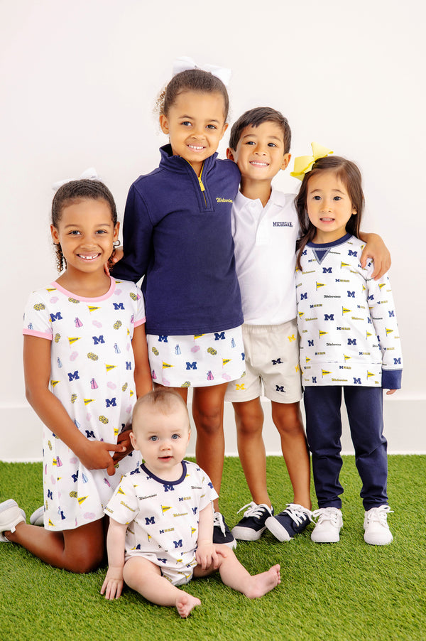 Five smiling children pose together on a green carpet, dressed in University of Michigan-themed outfits. Four are standing while one baby is seated in front. Some of the kids are sporting Wolverine gameday apparel, including The Beaufort Bonnet Company’s Michigan Hayword Half-Zip in Michigan Blue with Michigan Yellow and a University logo pullover. The background is a plain white wall.
