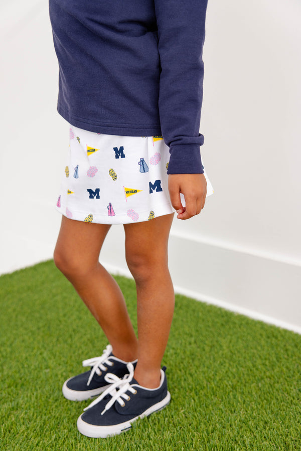 A child wearing navy sneakers, a navy long-sleeve shirt, and The Beaufort Bonnet Company’s Michigan Searcy Skort - Bold in Blue with Worth Avenue White, featuring University of Michigan-themed prints, stands on green artificial grass.