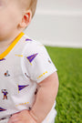 A baby wearing The Beaufort Bonnet Company’s LSU Short Sleeve Patton Play Bubble – Tenacious Tigers with LSU Gold sits on green grass. Only the baby’s left side, shoulder, and arm are visible.