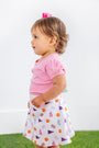 A young child with light brown hair, dressed in a pink shirt and The Beaufort Bonnet Company’s Clemson Searcy Skort - Tiger Triumph with Worth Avenue White, stands on green grass against a plain white background.