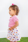 A young child with light brown hair, dressed in a pink shirt and The Beaufort Bonnet Company’s Clemson Searcy Skort - Tiger Triumph with Worth Avenue White, stands on green grass against a plain white background.