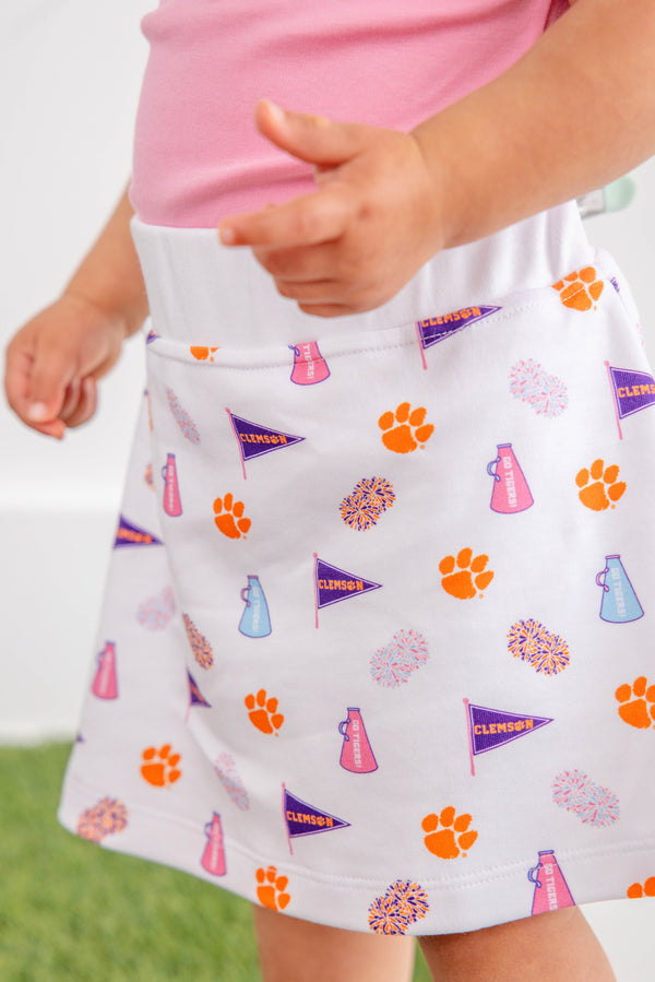A child is wearing The Beaufort Bonnet Company’s Clemson Searcy Skort – Tiger Triumph with Worth Avenue White, featuring orange paw prints, purple pennants, pink megaphones, and colorful pom-poms. The skort is paired with a light pink shirt, and the background is blurred.