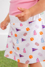 A child is wearing The Beaufort Bonnet Company’s Clemson Searcy Skort – Tiger Triumph with Worth Avenue White, featuring orange paw prints, purple pennants, pink megaphones, and colorful pom-poms. The skort is paired with a light pink shirt, and the background is blurred.
