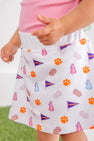 A child is wearing The Beaufort Bonnet Company’s Clemson Searcy Skort – Tiger Triumph with Worth Avenue White, featuring orange paw prints, purple pennants, pink megaphones, and colorful pom-poms. The skort is paired with a light pink shirt, and the background is blurred.