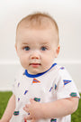 A baby with blue eyes and light brown hair wears The Beaufort Bonnet Company’s Florida Short Sleeve Patton Play Bubble – Florida Forever with Florida Blue, crafted from 100% Pima Cotton and adorned with small Florida Gators logos and flags in the playful Florida Forever print, while sitting against a white background and green floor.