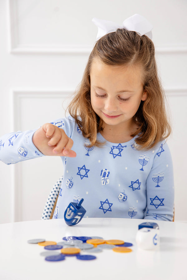 A young girl in The Beaufort Bonnet Company’s Long Sleeve Polly Play Dress – Eight Nights to Remember, adorned with festive Hanukkah symbols, smiles as she spins a dreidel at a white table with chocolate coins, looking down and wearing a white bow in her hair.