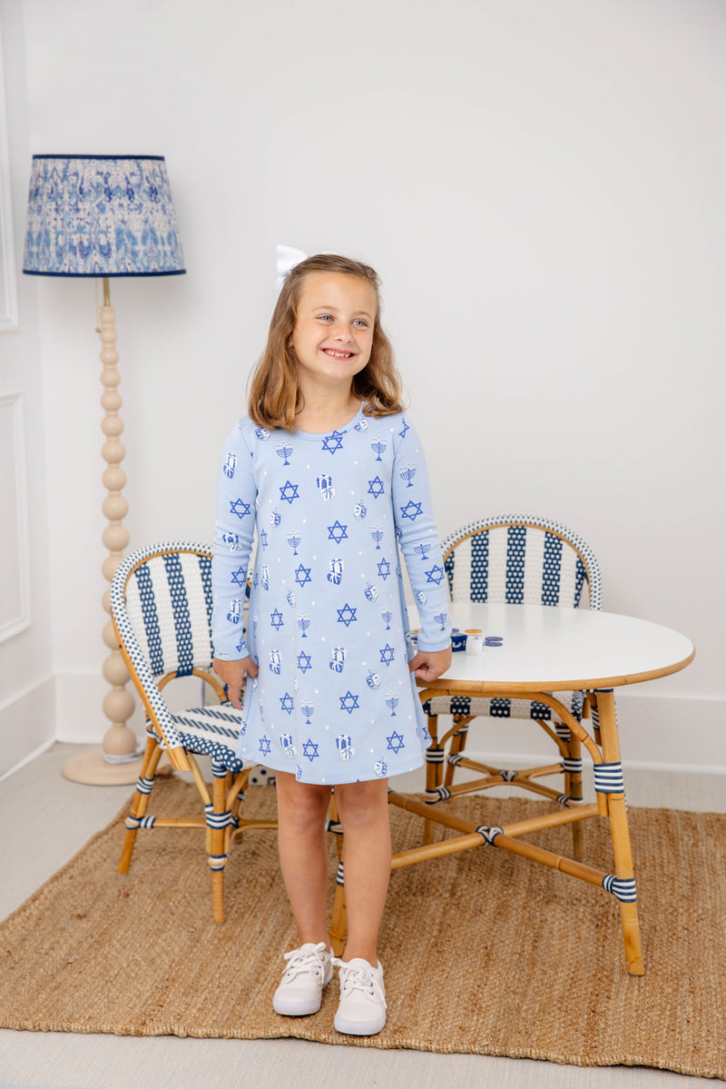 A smiling young girl wears The Beaufort Bonnet Company’s Long Sleeve Polly Play Dress - Eight Nights to Remember, crafted from soft Pima cotton with charming Star of David patterns. She stands in front of a small table and chairs, her look completed by a white hair bow and white sneakers, as a blue-patterned lamp glows in the background.