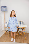 A smiling young girl wears The Beaufort Bonnet Company’s Long Sleeve Polly Play Dress - Eight Nights to Remember, crafted from soft Pima cotton with charming Star of David patterns. She stands in front of a small table and chairs, her look completed by a white hair bow and white sneakers, as a blue-patterned lamp glows in the background.