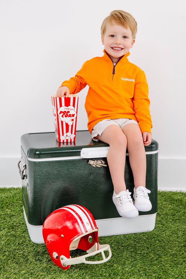 A young boy wearing The Beaufort Bonnet Company’s Clemson Hayword Half-Zip in Clemson Orange with Clemson Purple and Worth Avenue White sits on a green cooler, holding a striped popcorn box. A red football helmet rests on the artificial grass in front of him. He smiles, with a white wall as the backdrop.