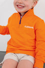 A smiling child sits with relaxed arms, wearing The Beaufort Bonnet Company’s Clemson Hayword Half-Zip in Clemson Orange with Clemson Regalia and Worth Avenue White, paired with light shorts; only the lower half of their face is visible.
