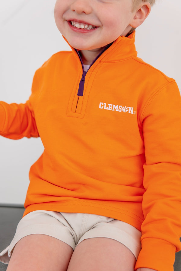 A smiling child sits with relaxed arms, wearing The Beaufort Bonnet Company’s Clemson Hayword Half-Zip in Clemson Orange with Clemson Regalia and Worth Avenue White, paired with light shorts; only the lower half of their face is visible.