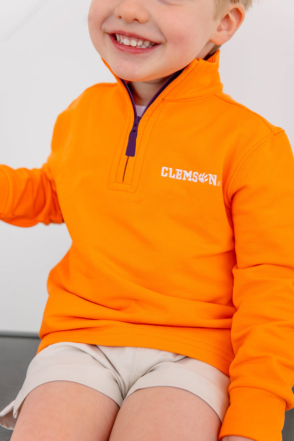 A smiling child wears The Beaufort Bonnet Company’s Clemson Hayword Half-Zip in Clemson Orange with Clemson Purple and Worth Avenue White, featuring an embroidered logo, paired with light-colored shorts and seated against a plain background—perfect for game day layering.