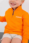 A smiling child wears The Beaufort Bonnet Company’s Clemson Hayword Half-Zip in Clemson Orange with Clemson Purple and Worth Avenue White, featuring an embroidered logo, paired with light-colored shorts and seated against a plain background—perfect for game day layering.