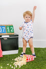 A toddler wearing The Beaufort Bonnet Company’s Ole Miss Short Sleeve Patton Play Bubble - Gawsh Almighty with Nantucket Navy stands on the grass next to spilled popcorn, raising one arm. Behind them are a cooler, number blocks, and a foam finger.