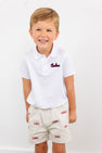 Smiling young boy wearing The Beaufort Bonnet Company’s South Carolina Prim & Proper Polo in Worth Avenue White, featuring an embroidered South Carolina logo, paired with beige shorts adorned with multiple red USC logos, standing against a plain white background.