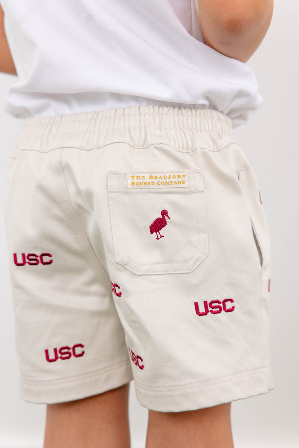 A child is wearing cream-colored South Carolina Critter Sheffield Shorts in Saratoga Stone by The Beaufort Bonnet Company, featuring the USC logo and a red embroidered bird, with “The Beaufort Bonnet Company” stitched above the back pocket.