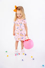 A young girl with a bright orange bow in her hair wears the charming Polly Play Dress - Candy & Critters (Sandpearl Pink) by The Beaufort Bonnet Company, crafted from soft Pima Cotton and adorned with whimsical pumpkins and cats. She holds a pink pumpkin bucket and stands among scattered colorful candies on a white background, making this girls’ dress perfect for festive occasions.